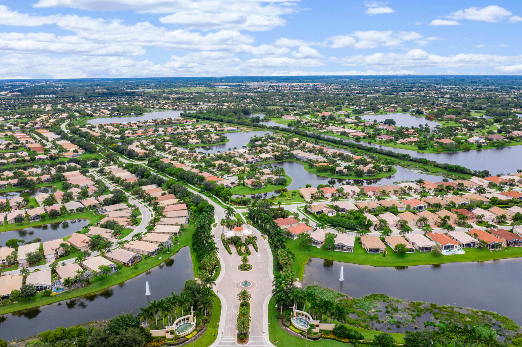 Aerial view of a suburban neighborhood by Akel Homes, one of the Best Home Builders in Florida, featuring rows of houses, streets, lakes, green spaces, and a gated entrance lined with palm trees.
