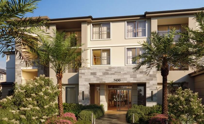 Three-story modern apartment building, Mosaic Palms, features a stone and stucco exterior, large windows, and a landscaped entrance with palm trees and shrubs. Now pre-leasing in Lake Worth FL.