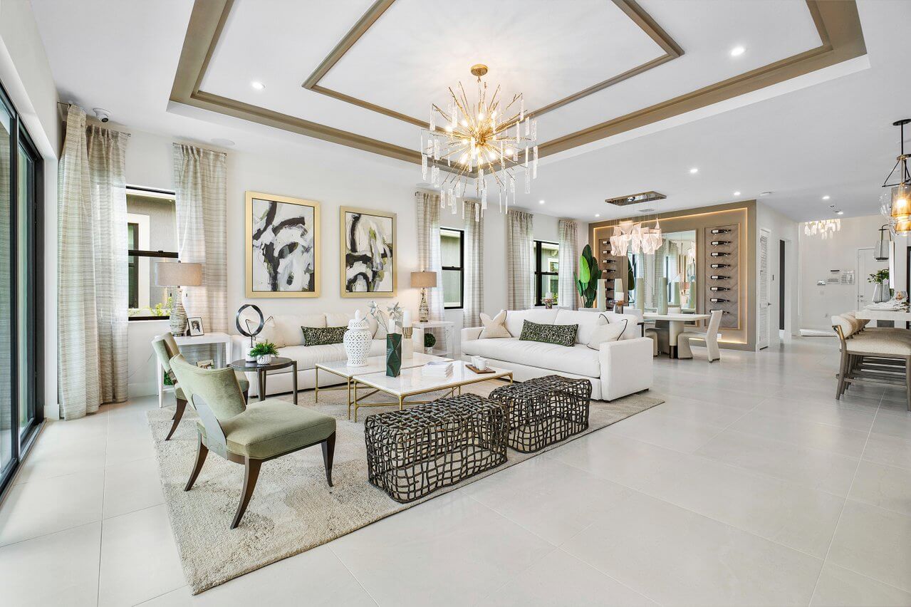 Modern open-concept living and dining area with white sofas, green chairs, abstract art, chandeliers, large windows, and light gray tile flooring. This beautifully furnished model home at Solana Bay is now for sale.