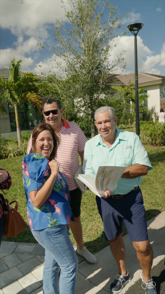 Three adults stand outside on a sunny day, smiling at the camera during a grand opening. One holds a book or binder. Palm trees and buildings can be seen in the background.