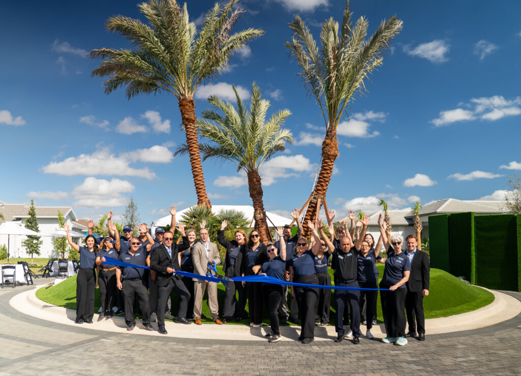 A group of people stands outdoors in front of palm trees, holding a large blue ribbon at a ribbon-cutting ceremony, with several people raising their hands and smiling.