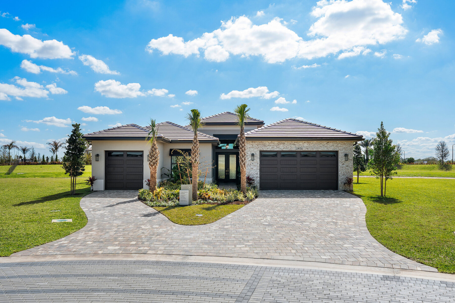 Modern single-story house with three-car garage, palm trees, and landscaped front yard, set under a blue sky with scattered clouds—one of 18 Model Homes designed for luxurious, comfortable living.
