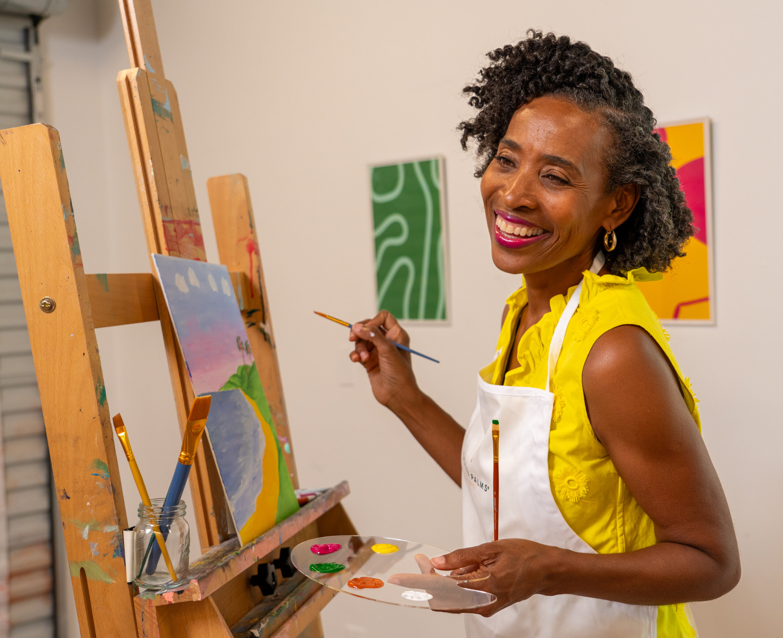 A woman in a yellow top and white apron smiles while painting at an easel in an art studio, holding a palette and brush—imagine your days inspired by creativity at Catalina Palms.