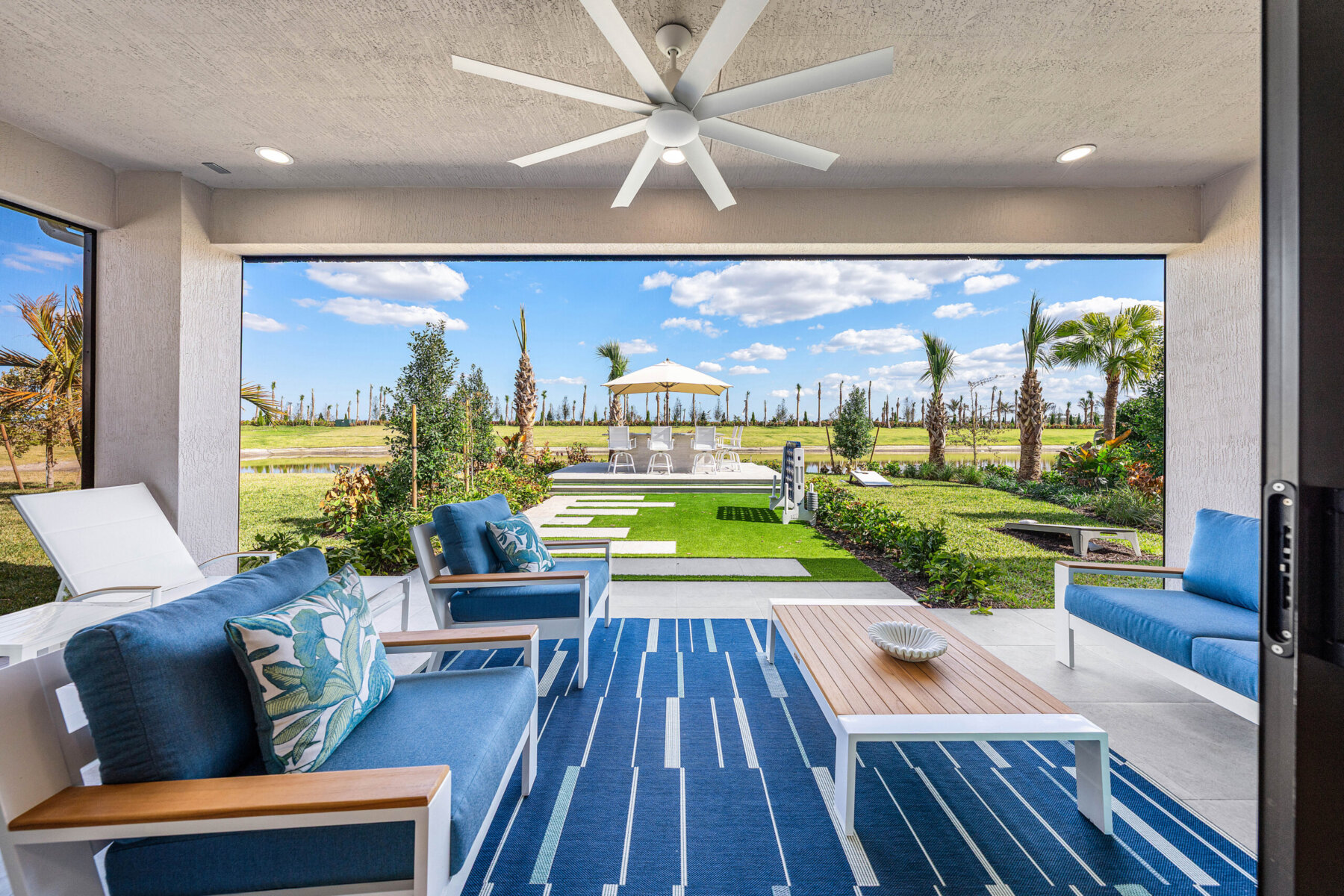 Covered patio with blue and white outdoor furniture, ceiling fan, and a view of a landscaped backyard with pool and lounge chairs—enjoy resort-style living for less while adding value to your home.