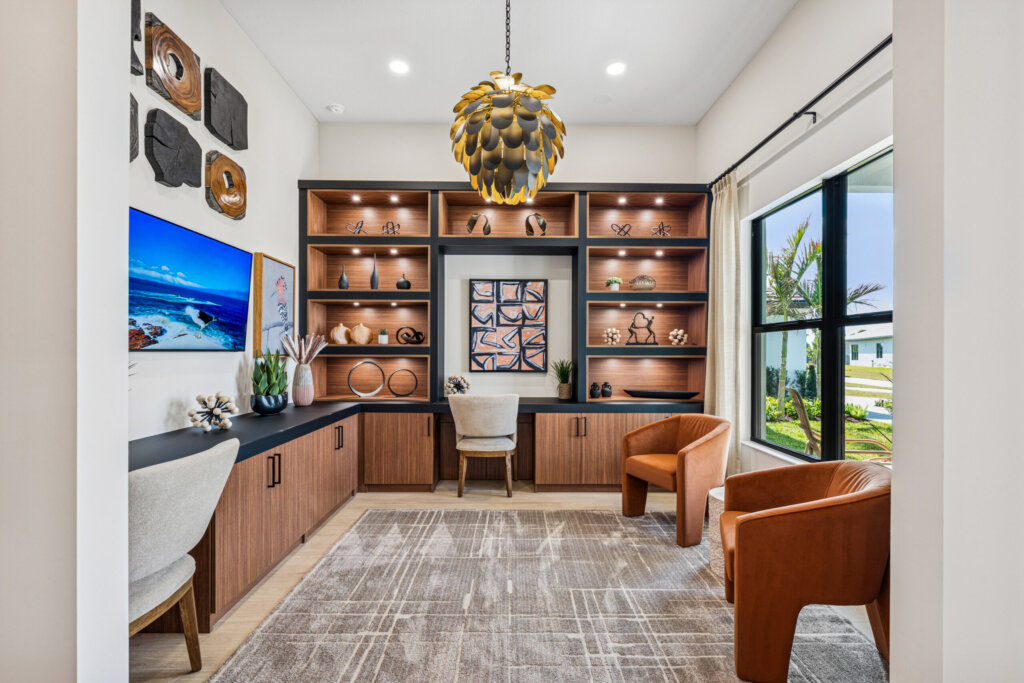 Modern home office with built-in wooden shelves, two tan chairs, two gray desk chairs, a large rug, decorative objects, framed art, and a window overlooking houses and Catalina Palms—perfect for a stylish home lifestyle.