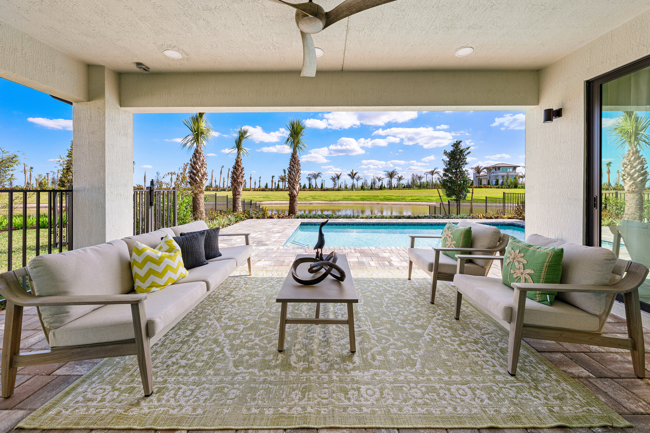 Covered patio with two sofas, a coffee table, and a green patterned rug overlooking a swimming pool, palm trees, and a landscaped yard under a blue sky—a relaxing oasis in the heart of Catalina Palms.