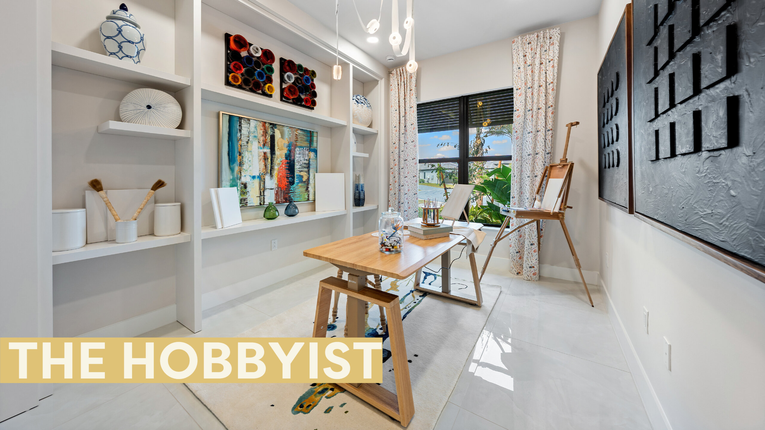A bright art studio in Catalina Palms with shelves holding decor, a table with art supplies, an easel by the window, and large black wall art; text reads "THE HOBBYIST." Perfectly fit for a creative home.