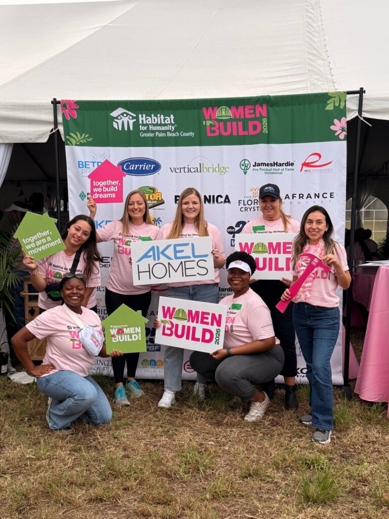 Eight women in pink “Women Build” shirts pose in front of a sponsor banner at a Habitat for Humanity event, holding signs and smiling for the photo during Women Build 26.