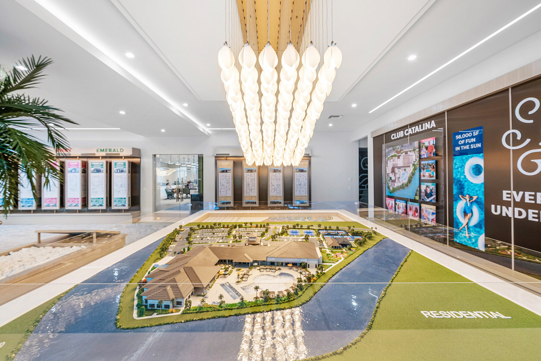 A detailed architectural scale model of Catalina Palms residential development is displayed indoors under modern pendant lights, with informational posters on the walls in the background, inviting curiosity for a first visit.