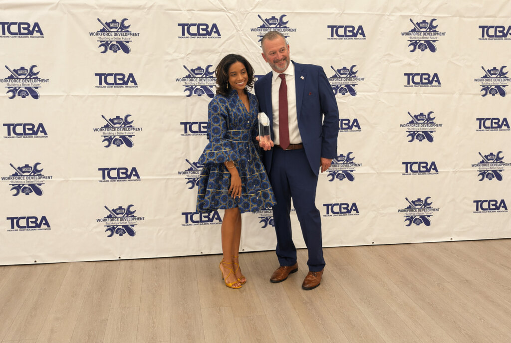 Two people stand posing for a photo at the Parade of Homes event, with one holding a trophy. A branded step-and-repeat banner is in the background.