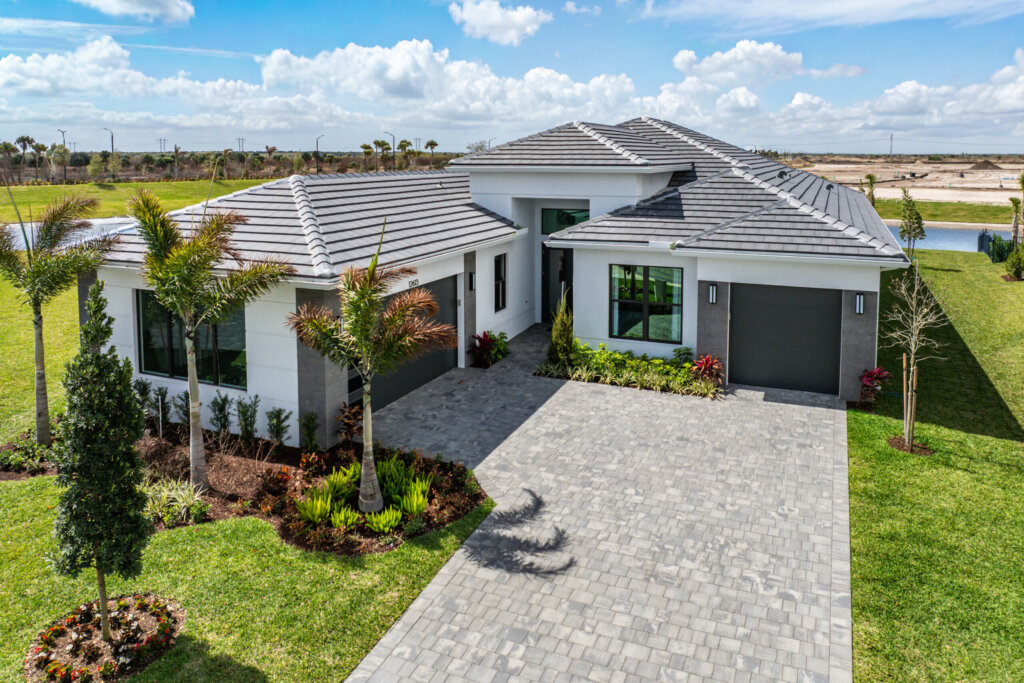 Modern single-story house with gray tile roof, two garages, palm trees, landscaped yard, and a wide stone driveway under a partly cloudy sky—explore it all in Akel Homes video tours.
