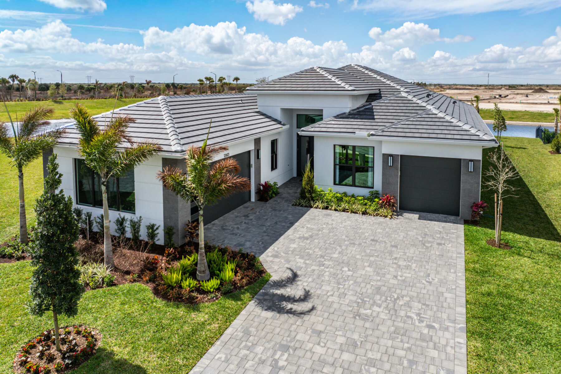 Modern single-story house with gray tile roof, two garages, palm trees, landscaped yard, and a wide stone driveway under a partly cloudy sky—explore it all in Akel Homes video tours.