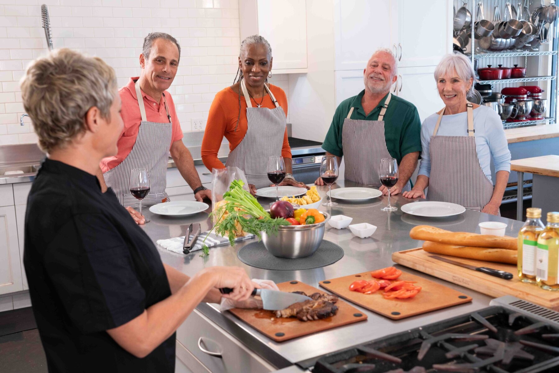 A chef prepares food at a kitchen counter in a lively culinary lab while four adults in aprons watch, with vegetables, bread, and wine glasses on display.