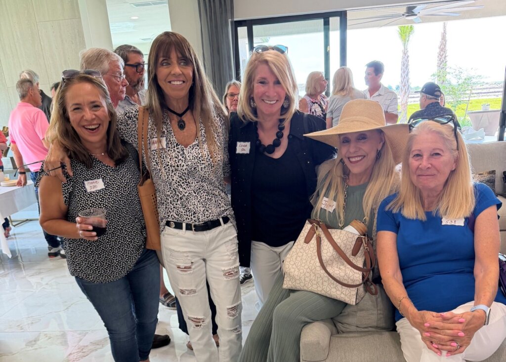 A group of five women, some standing and some sitting, pose and smile at a Lifestyle event during the Homebuyer Happy Hour at Catalina Palms; other attendees are visible in the background.