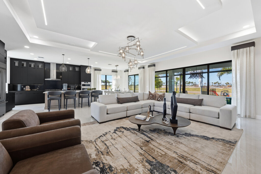 Spacious modern living room with beige sofas, brown armchairs, a large area rug, and an open kitchen with more black cabinets and bar stools in the background. Large windows provide outdoor views.