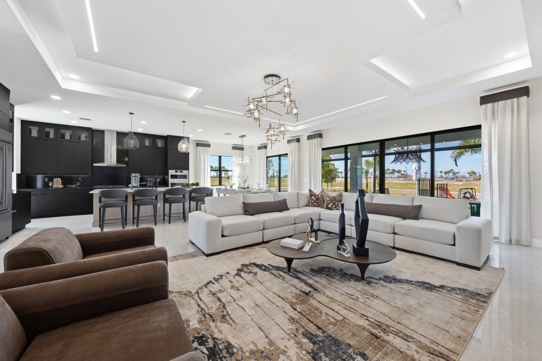 Spacious modern living room with beige sofas, brown armchairs, a large area rug, and an open kitchen with more black cabinets and bar stools in the background. Large windows provide outdoor views.