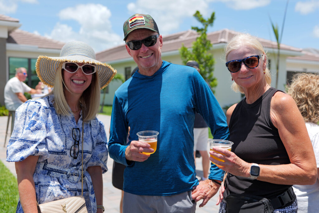 Three adults wearing sunglasses and casual summer clothing stand outdoors, smiling and holding drinks at a Club Catalina groundbreaking celebration during the day.