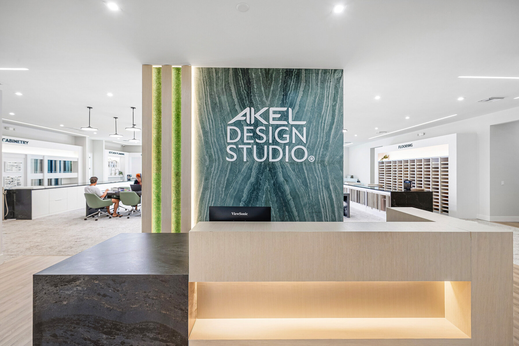 Reception area of Akel Design Studio featuring a stylish front desk, illuminated signage, modern decor, and a vibrant workspace with people seated at tables in the background.