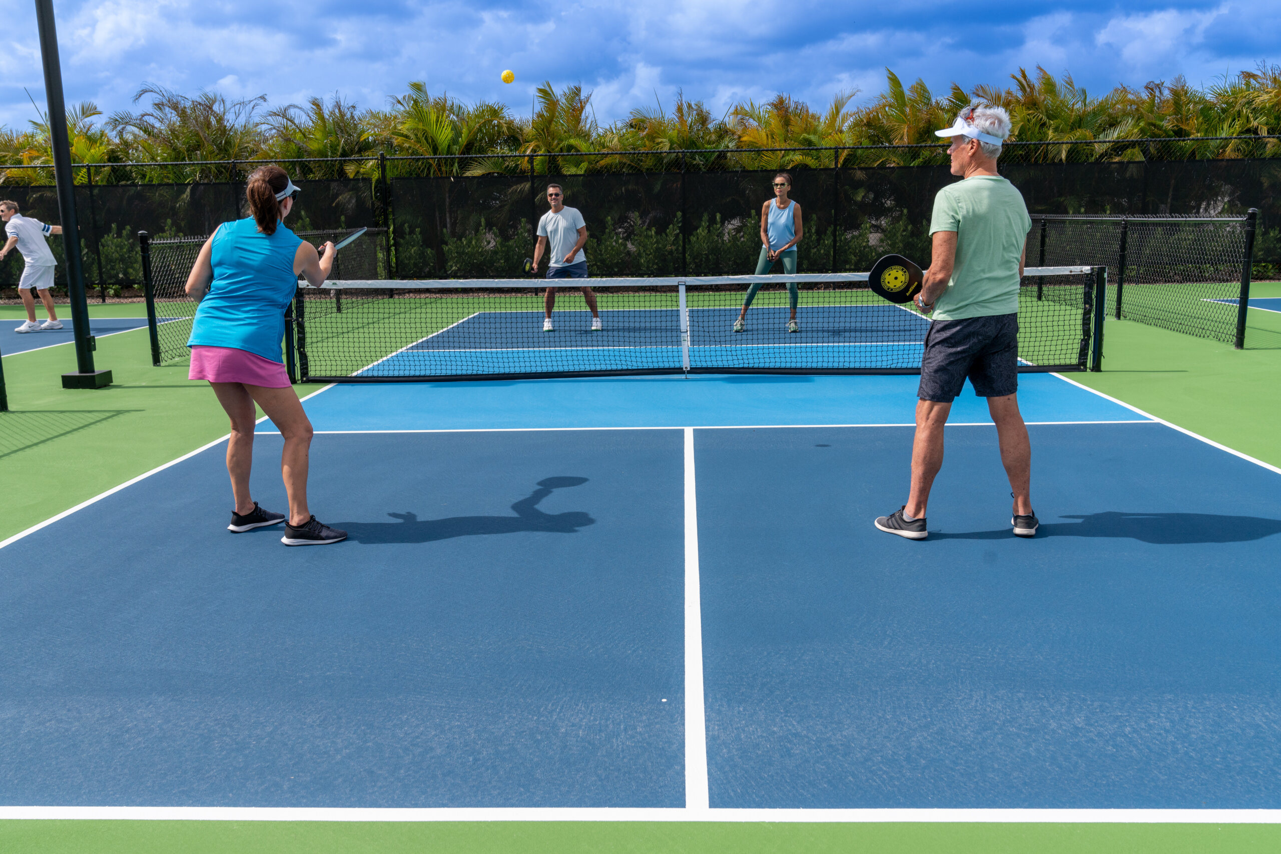 Four adults play pickleball on an outdoor court at Sundance Vitality Village, with two players on each side of the net under a partly cloudy sky.