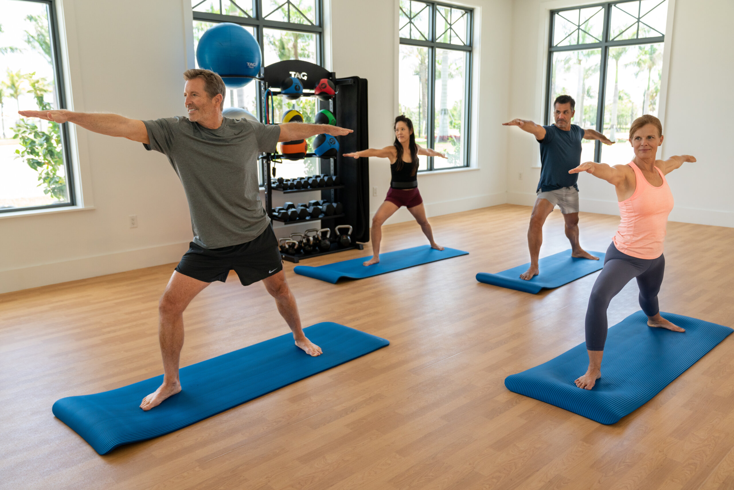 Four adults practice yoga in a bright studio at Vitality Village, standing on blue mats in a warrior pose. Exercise equipment is visible in the background, adding to the vibrant Sundance energy of the session.