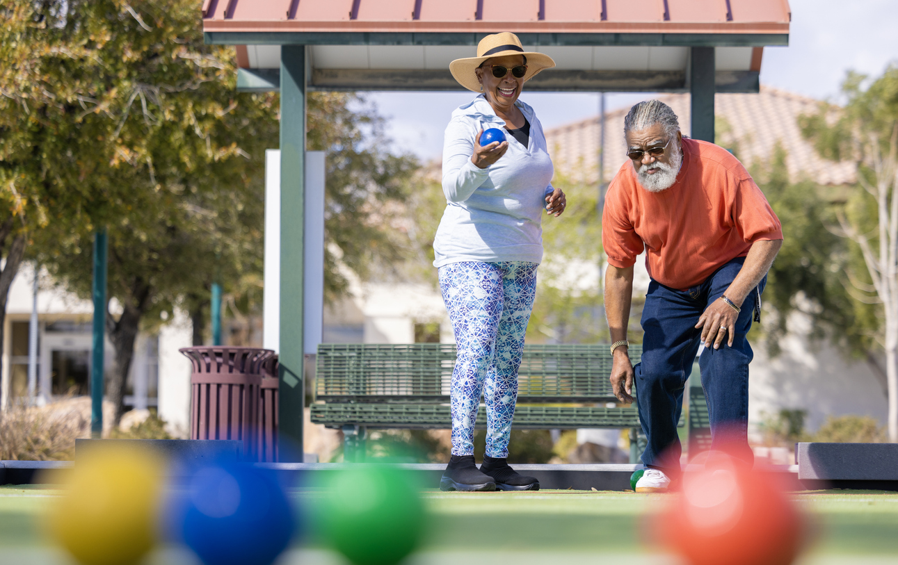 Two seniors play bocce ball outdoors at Sundance Vitality Village. The woman smiles while holding a blue ball, and the man leans forward as colored balls rest on the green court.