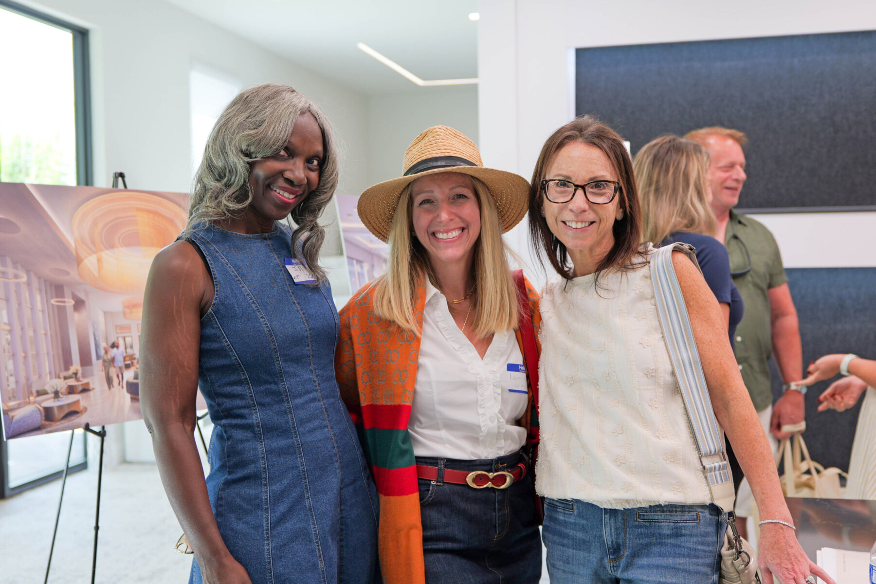 Three women smiling at an event, with people and architectural renderings in the background.