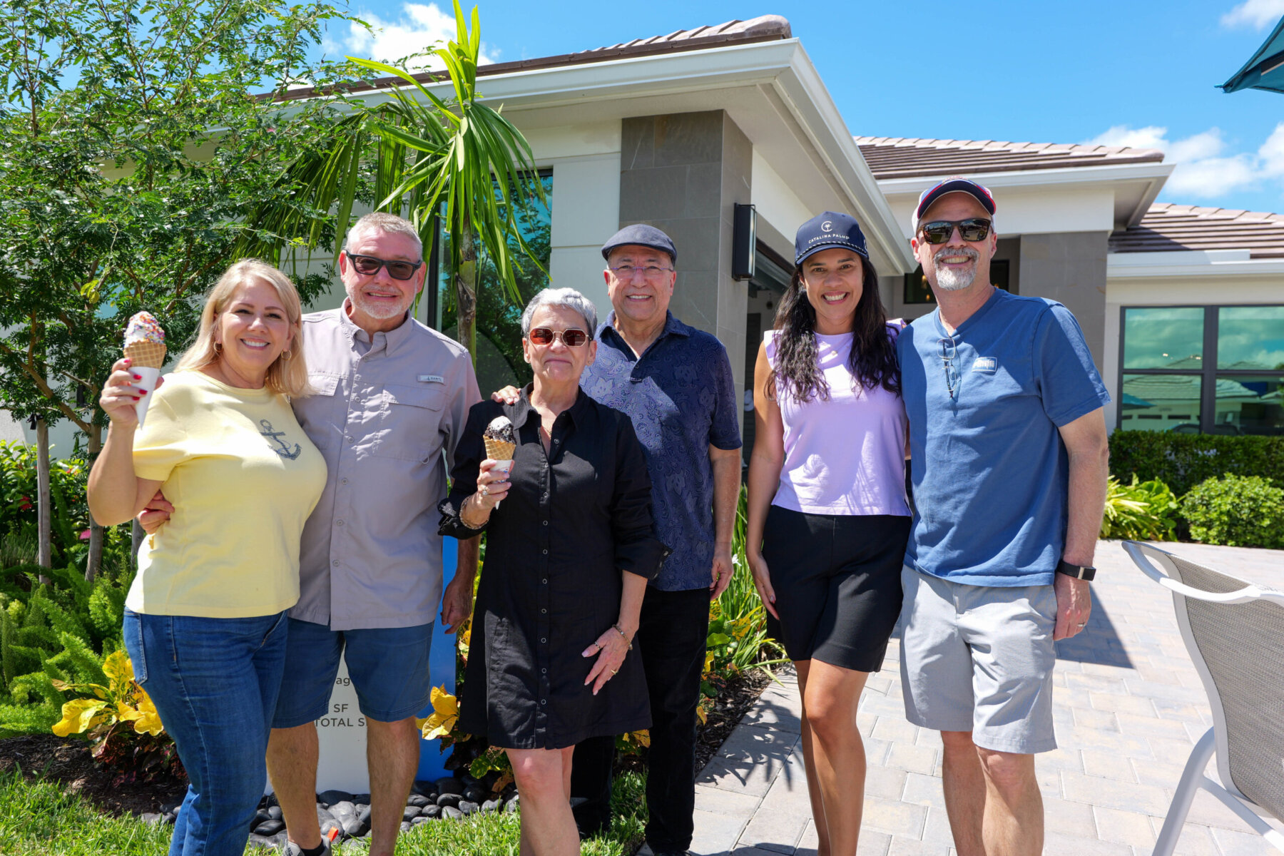 Six adults stand outdoors on a sunny day, smiling and holding ice cream cones in front of a building with greenery in the background.