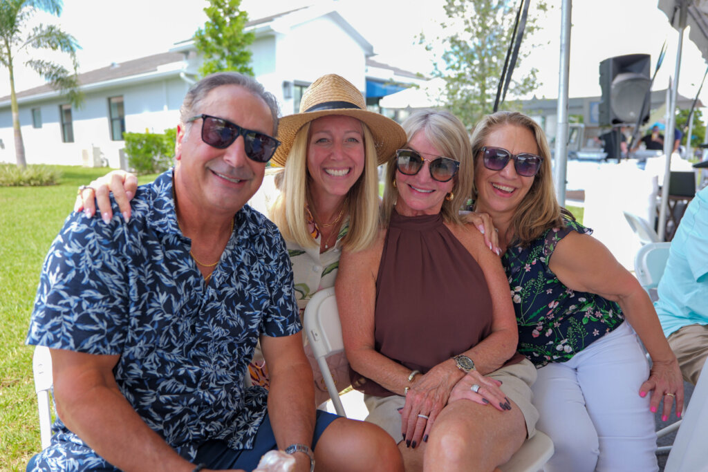 Four adults in summer clothes and sunglasses smile outdoors at Catalina Palms, with a house, grass, and trees in the background.