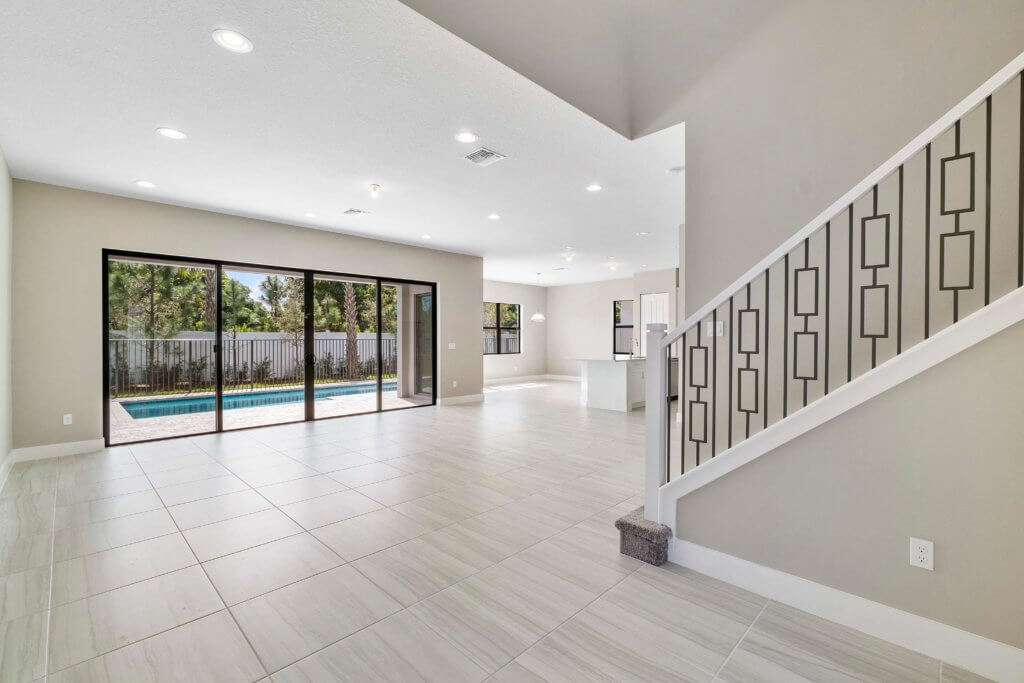 A spacious, unfurnished living room in Villamar at Toscana Isles features light-colored tile flooring. Large sliding glass doors lead to an outdoor pool area. The interior boasts neutral-colored walls and a stairway with a modern railing design, perfect for contemporary living.