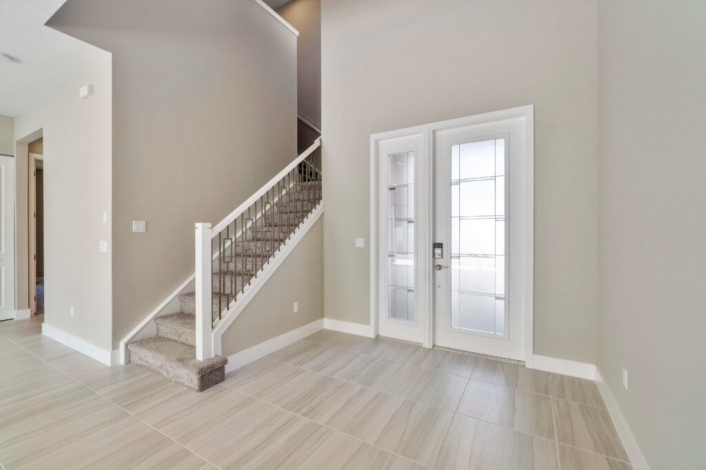 A modern interior at Villamar at Toscana Isles features a staircase with carpeted steps on the left and elegant glass double doors on the right. The floor is tastefully tiled, and neutral-toned walls complete this sophisticated space.