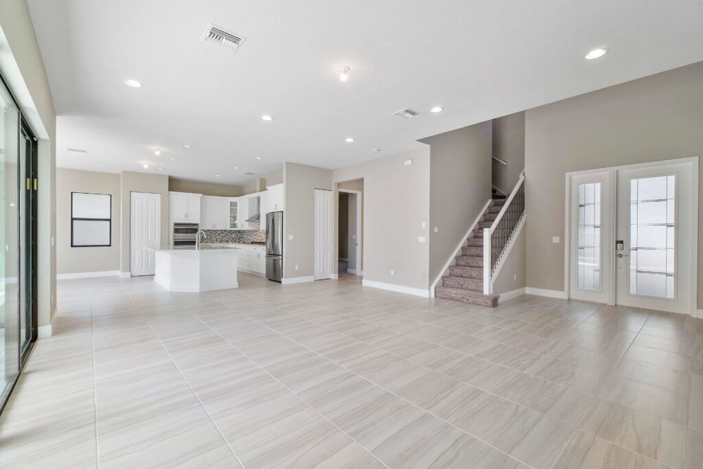 The spacious interior of this Villamar at Toscana Isles home boasts a modern, unfurnished design with light-colored tile flooring. It features an open-plan kitchen with white cabinets and a center island, adjacent to a living area. A staircase leads to the upper floor, where elegant glass doors are visible.