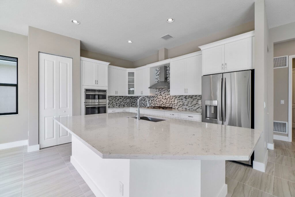 A modern kitchen at Villamar at Toscana Isles features white cabinets, stainless steel appliances, and a large quartz island with a built-in sink. The backsplash boasts a geometric tile design, while recessed lighting beautifully brightens the space.