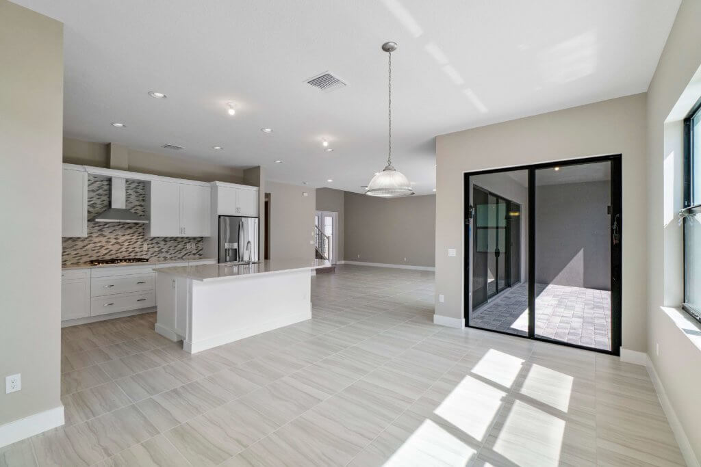 A spacious, unfurnished modern kitchen and living area at Villamar at Toscana Isles boasts light-colored tile flooring. The kitchen features white cabinets, a large island, and a stainless steel refrigerator. Large glass doors lead to an outdoor patio, filling the space with bright natural light.