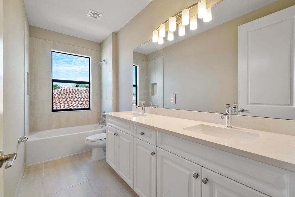 In a modern bathroom at Villamar at Toscana Isles, a large mirror graces the double sink vanity with white cabinets. The space is bathed in light from overhead fixtures and features a bathtub with shower. A window offers views of the tiled roof against a blue sky, creating an airy ambiance.