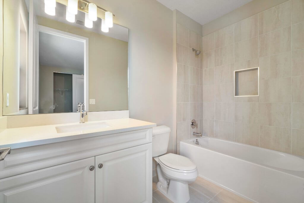A modern bathroom at Villamar at Toscana Isles features a sleek white vanity with a sink and a large mirror above. The room includes a toilet and a bathtub with a tiled wall. Bright lighting enhances the light neutral-toned walls, creating an inviting ambiance.