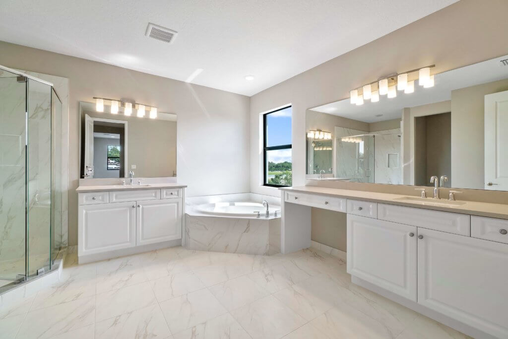 A spacious bathroom at Villamar at Toscana Isles boasts white cabinetry and light marble flooring. It features a double vanity with rectangular mirrors, a corner bathtub, and a glass-enclosed shower. A window allows natural light to stream in while offering serene views outside.