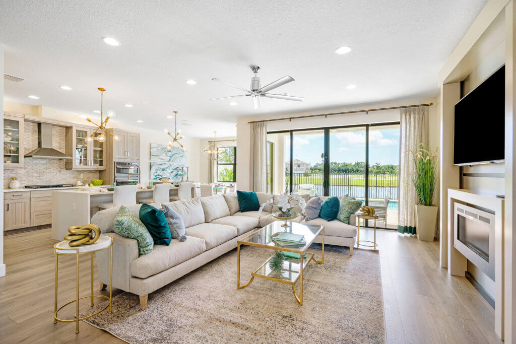 A spacious modern living room at Villamar at Toscana Isles features a beige sectional sofa adorned with assorted blue and green pillows. A large wall-mounted TV sits above an electric fireplace, while sliding glass doors reveal a pool view. The kitchen is visible in the background.