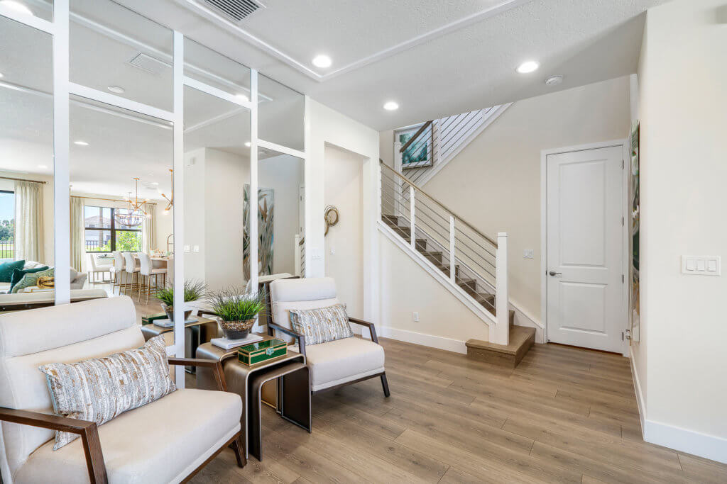 The modern living area at Villamar at Toscana Isles features two white cushioned chairs with decorative pillows, a small table with a plant, and mirrored panels on the wall. A staircase leads to the upper floor, while wooden flooring offers a peek into the inviting dining area.