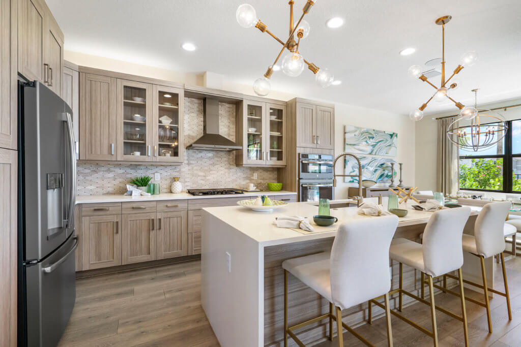 A modern kitchen at Villamar at Toscana Isles features light wood cabinets, marble countertops, and a large island with four upholstered bar stools. Two pendant lights hang above the island. Stainless steel appliances and a decorative backsplash enhance the space, while a window invites sunlight in.