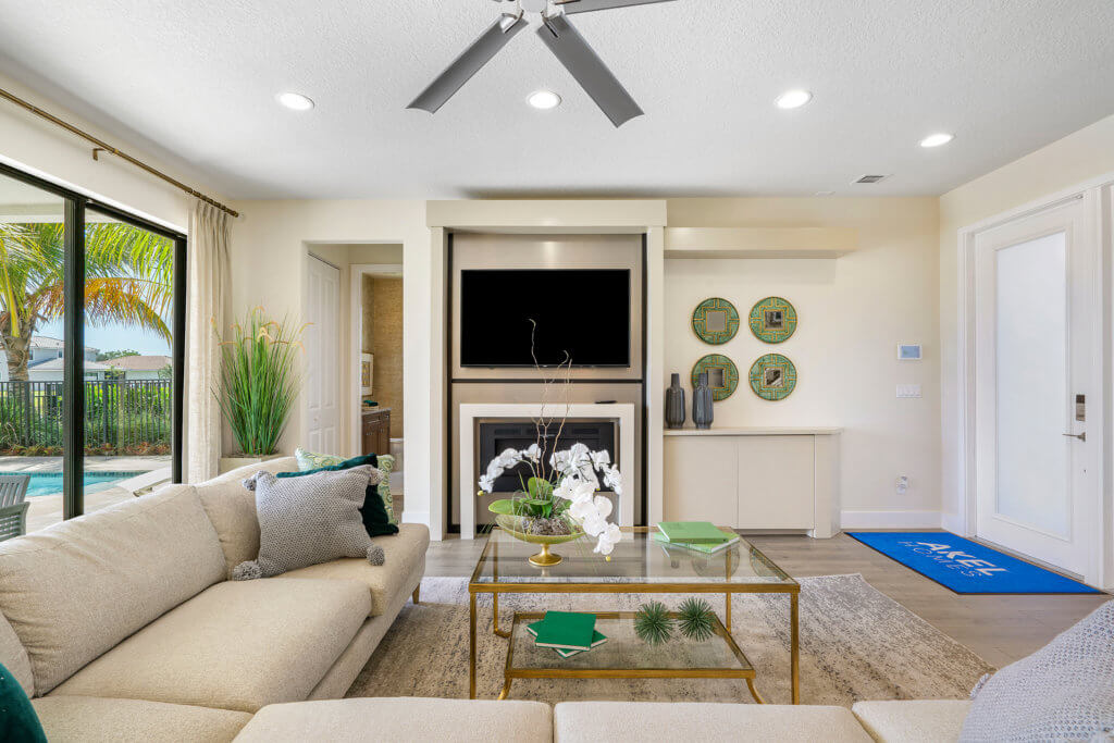 A modern living room at Villamar at Toscana Isles features a beige sectional sofa, glass coffee table, and fireplace. The flat-screen TV is mounted above the fireplace. Large windows with curtains open to a view of a pool and palm trees, complemented by a ceiling fan and recessed lights.
