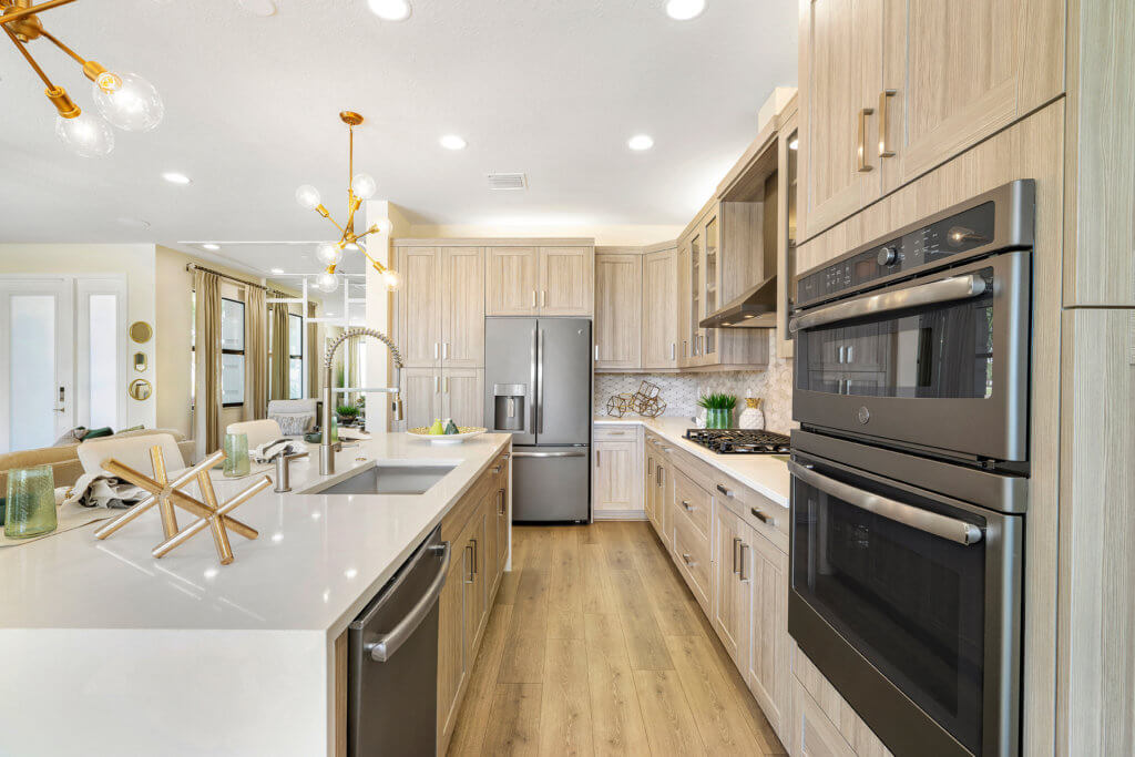 Discover the modern kitchen at Villamar at Toscana Isles, featuring light wood cabinets and dual built-in ovens complemented by a stainless steel refrigerator. A sleek white island with a sink and dishwasher takes center stage, illuminated by pendant lights. The dining area is elegantly visible in the background.