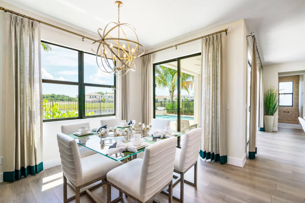 A bright dining room at Villamar at Toscana Isles features a glass table set for four. Large windows with light curtains showcase the backyard with palm trees and a pool. The modern chandelier illuminates the light wood floor, while a plant adds greenery to the corner.