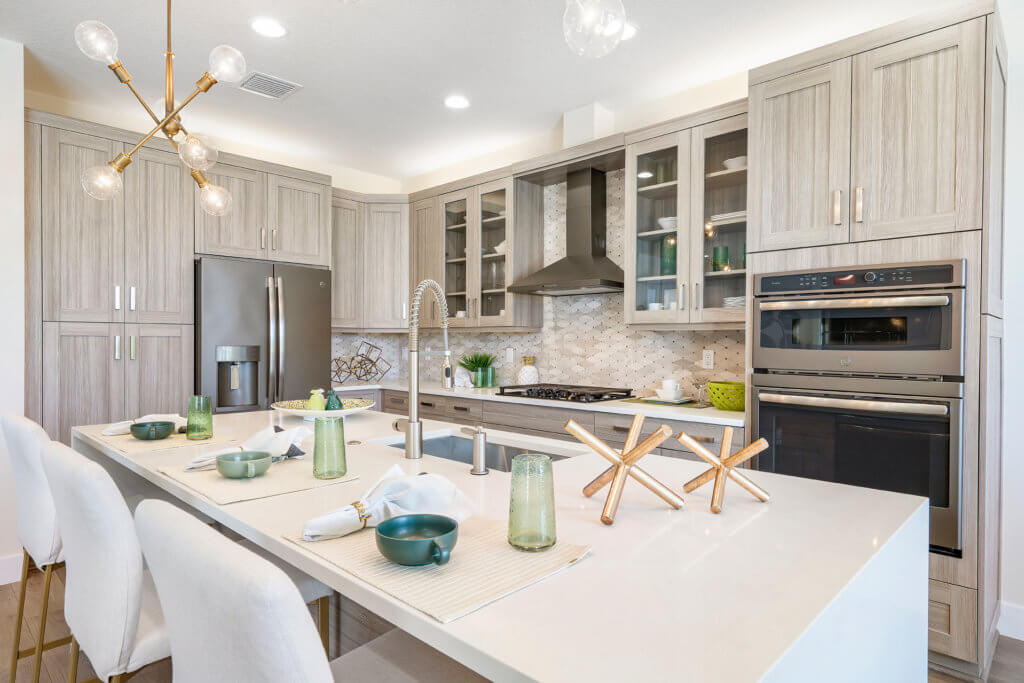 Located in Villamar at Toscana Isles, this modern kitchen boasts light wood cabinets, stainless steel appliances, and a sleek white island countertop. Four white chairs line the island, set with green glasses and dishes. A contemporary light fixture and geometric sculptures complete the decor.
