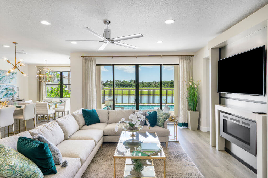 A modern living room in Villamar at Toscana Isles features a large beige sectional sofa with decorative pillows and a glass coffee table. The flat-screen TV is mounted above the fireplace. Expansive windows reveal the pool and scenic views, while a nearby dining area completes the space.