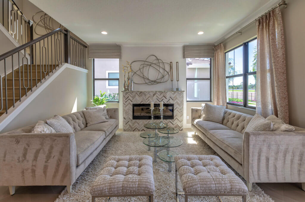A modern Villamar at Toscana Isles living room showcases two beige sofas facing each other, a geometric glass coffee table, and tufted ottomans atop a plush rug. A decorative sculpture adorns the herringbone-patterned fireplace, while large windows flood the space with natural light.