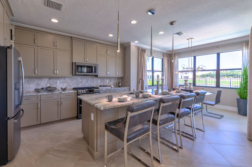 Spacious modern kitchen with beige cabinets and stainless steel appliances. A marble island has four high-backed stools, and decorative pendant lights hang above. A large window brightens the space, offering a glimpse of Villamar at Toscana Isles' lush greenery outside.