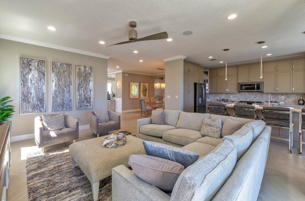 A contemporary living room in Villamar at Toscana Isles showcases a large L-shaped sofa, a central ottoman, and two armchairs. The space is adorned with abstract wall art and features an open-plan kitchen with bar stools and recessed lighting in the background.