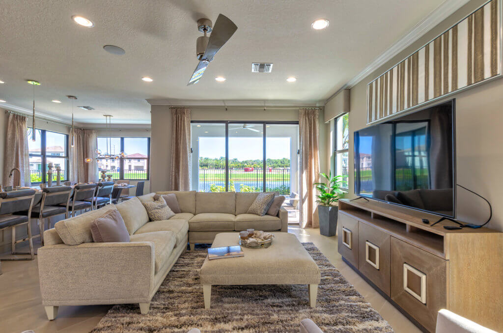 A modern living room at Villamar at Toscana Isles features a large beige sectional sofa with a matching ottoman on a textured rug. A flat-screen TV rests on a wooden console, while sliding glass doors reveal the patio and lush greenery outside.