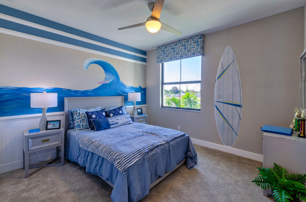 A bedroom at Villamar at Toscana Isles embraces a beach theme with blue striped bedding, a surfboard wall decoration, and a large wave mural. Two nightstands with lamps flank the bed, while a window offers views of greenery and sky. A ceiling fan gently stirs the coastal breeze above.