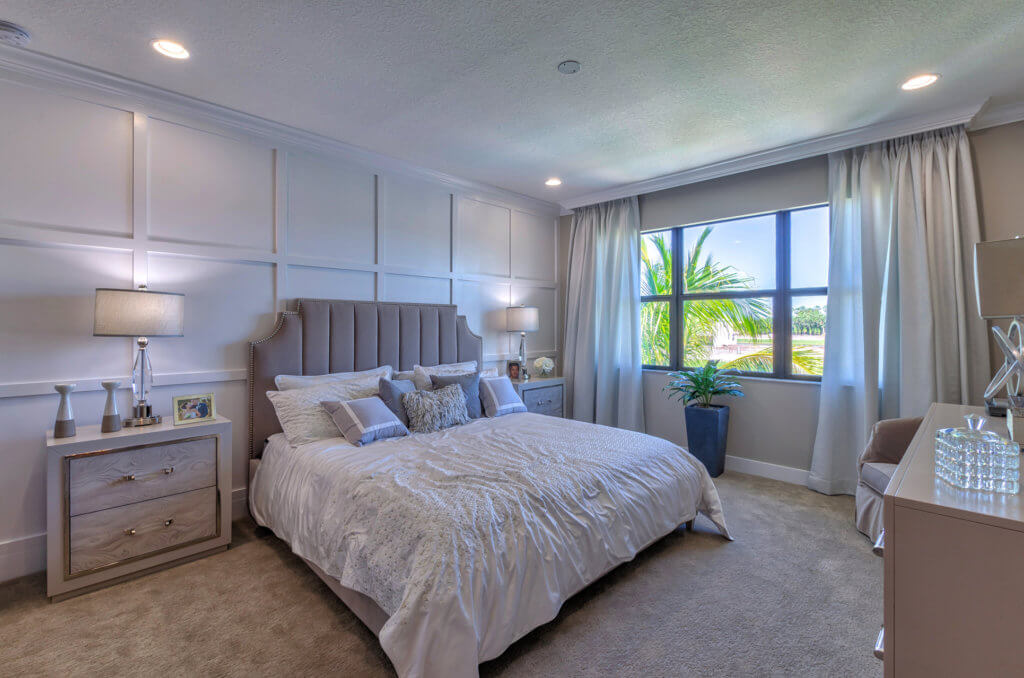 The bright bedroom at Villamar at Toscana Isles boasts a large bed with crisp white bedding and plush pillows. It features two bedside tables with lamps, a window draped in curtains framing lush greenery, and a potted plant by the window. The room is complete with carpeted floors and paneled wall decor.